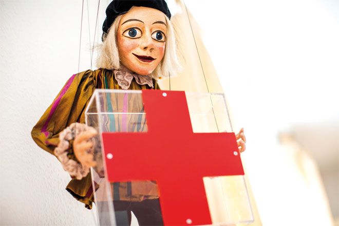 Foto: Eine Marionette hält vor sich ein großes, rotes Kreuz in den Händen.